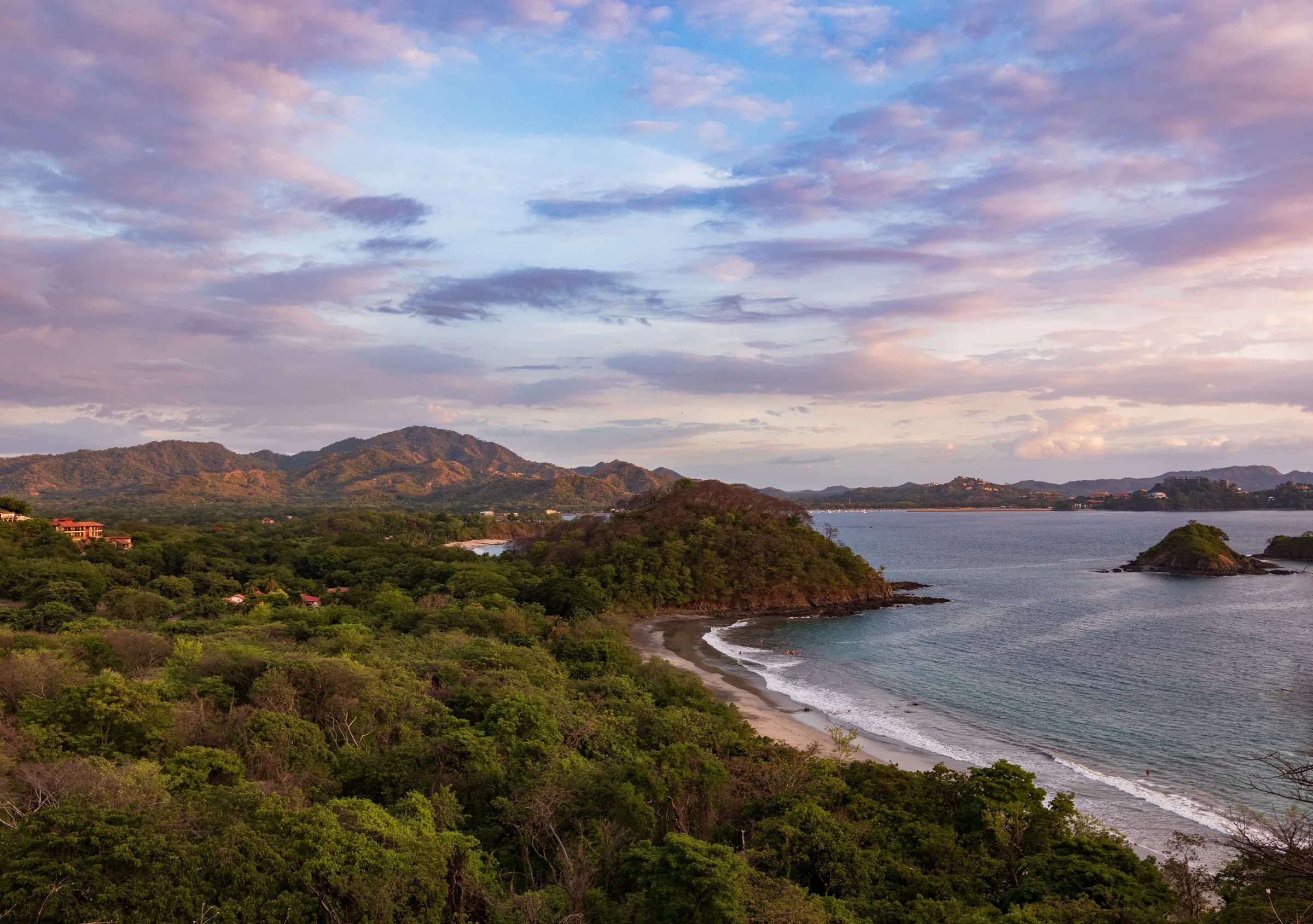 A forested headland, a sandy beach and a small rocky islet on the Guanacaste coast at dusk, with hills behind and pink clouds above.