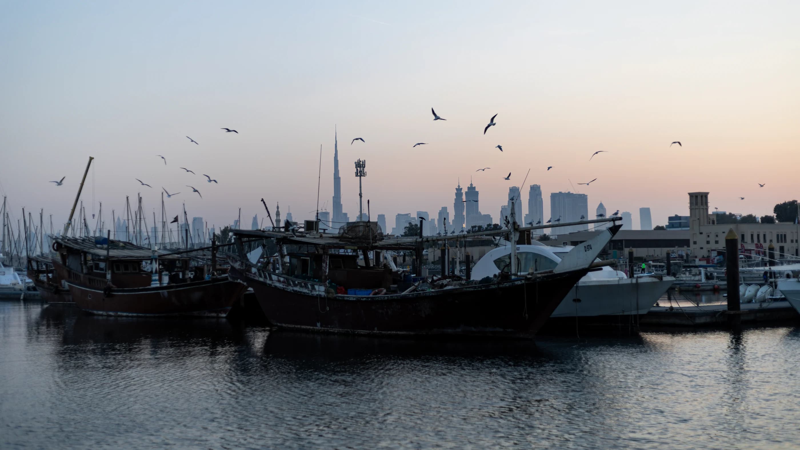 Wooden dhows in Umm Suqeim harbour at dawn, with gulls overhead and the Burj Khalifa on the Dubai skyline behind.
