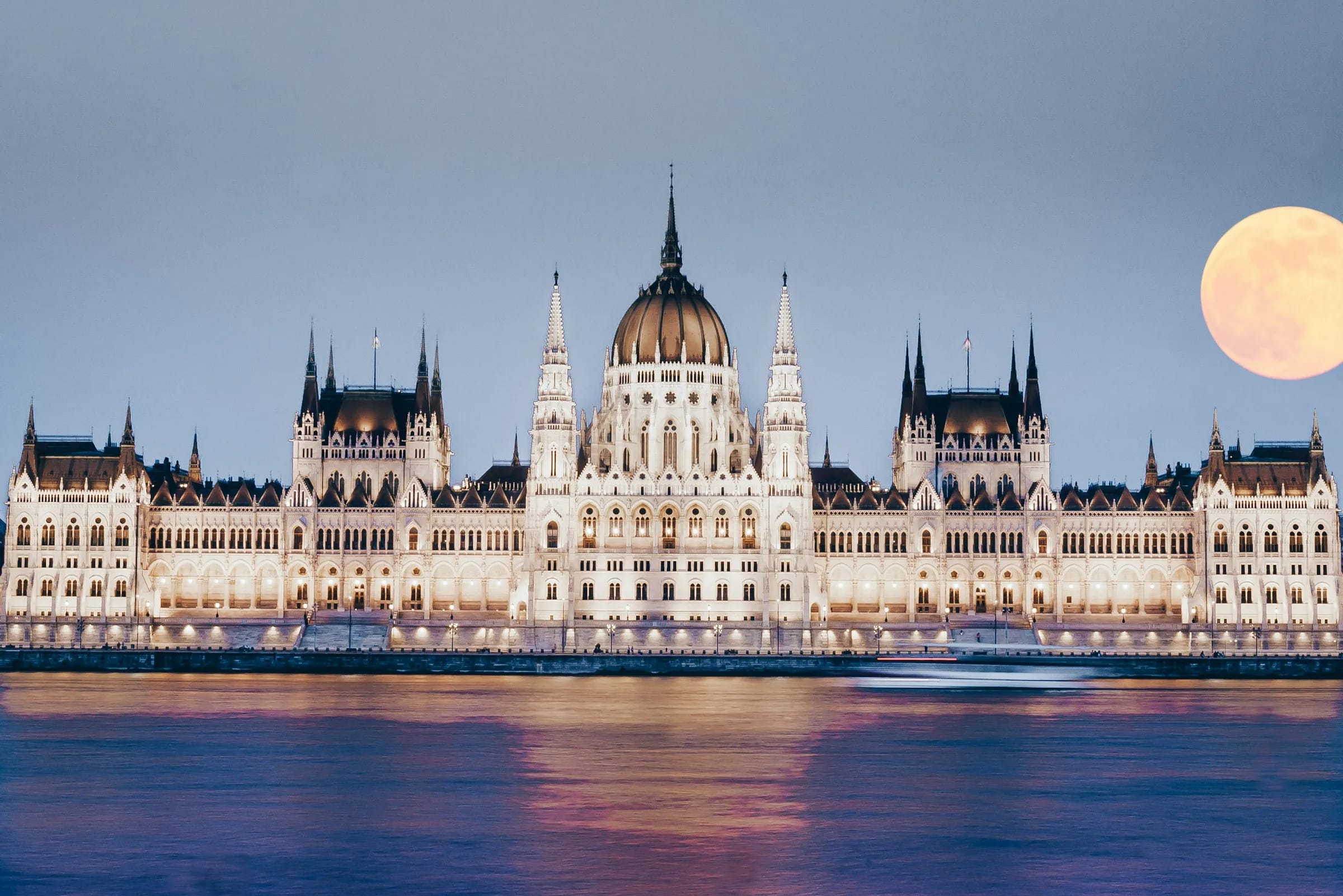 Das ungarische Parlamentsgebäude in der Abenddämmerung beleuchtet am Ufer der Donau in Budapest, daneben ein aufgehender Vollmond.