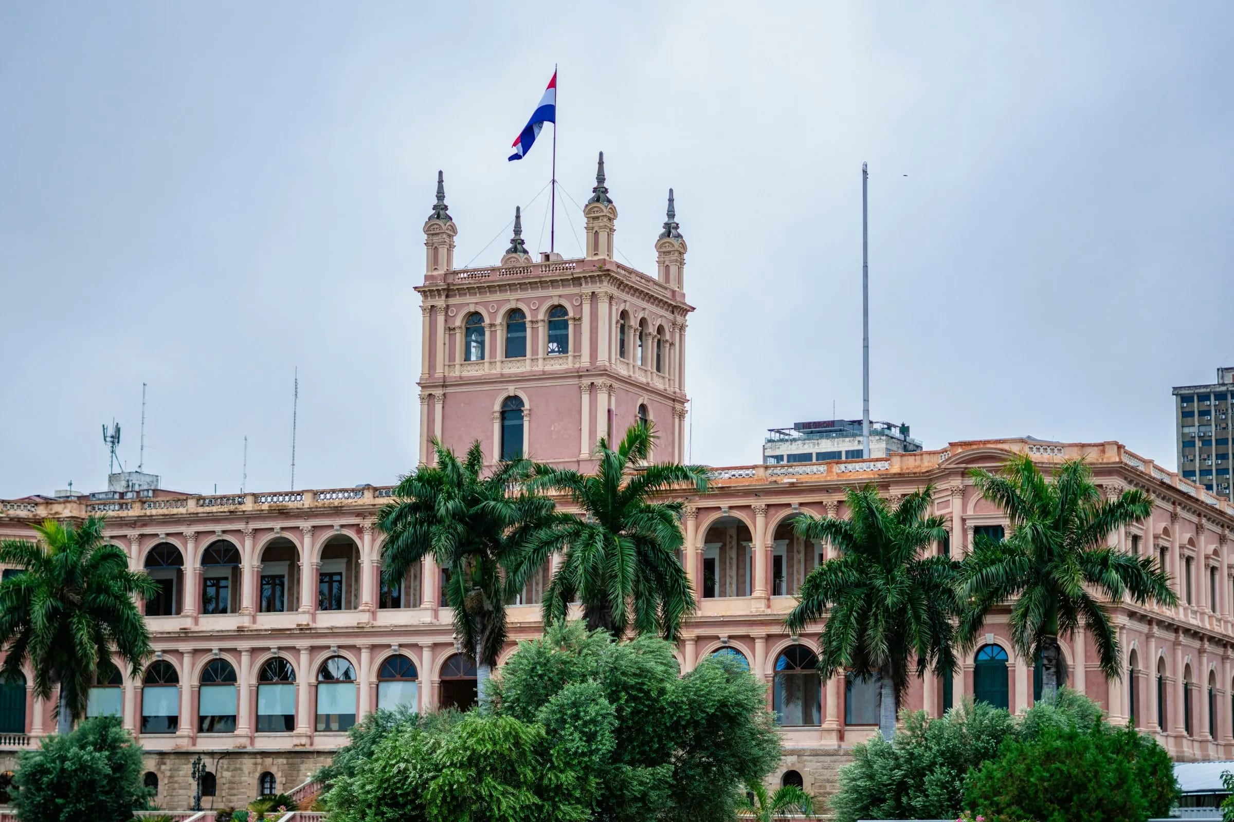 Ein rosafarbenes Regierungsgebäude im Kolonialstil mit einem Turm und Palmen davor, auf dem eine rot-weiß-blaue Flagge weht, in Asunción.