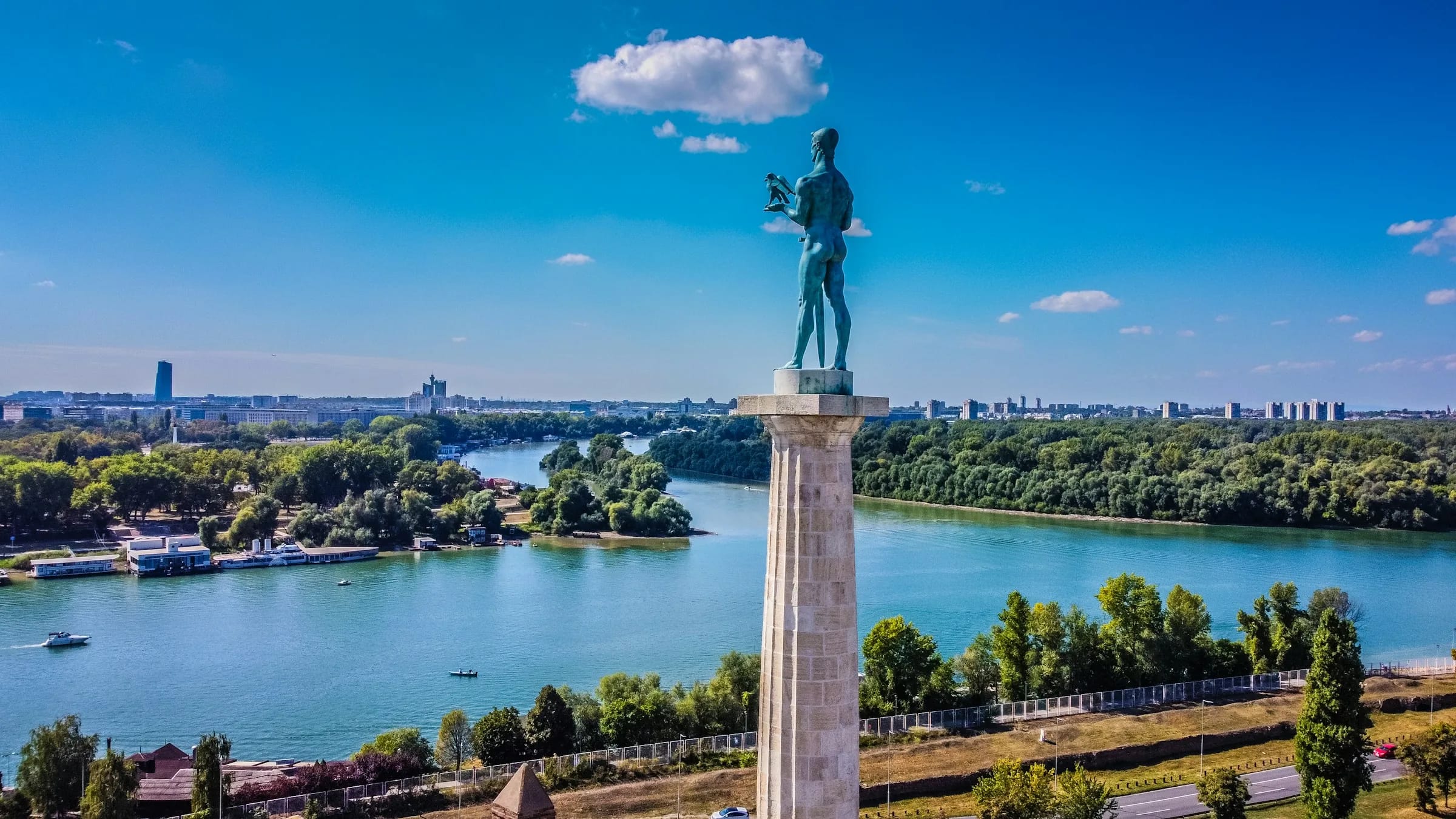 The Victor monument, a bronze figure on a stone column at Belgrade Fortress, above a wide river with wooded banks.