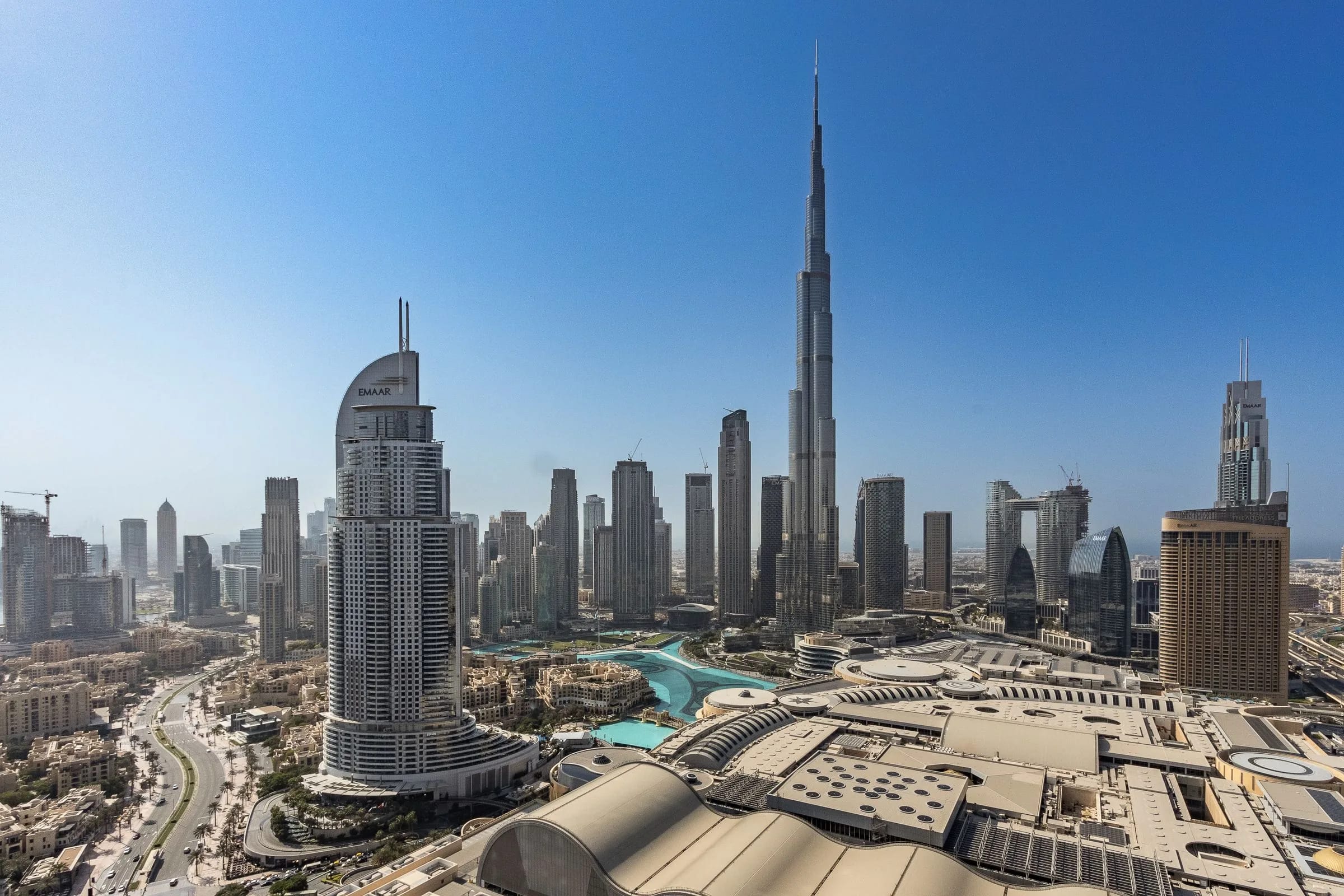 Luftaufnahme von Downtown Dubai mit dem Burj Khalifa über der Skyline und den Dächern der Dubai Mall unter klarem blauem Himmel.