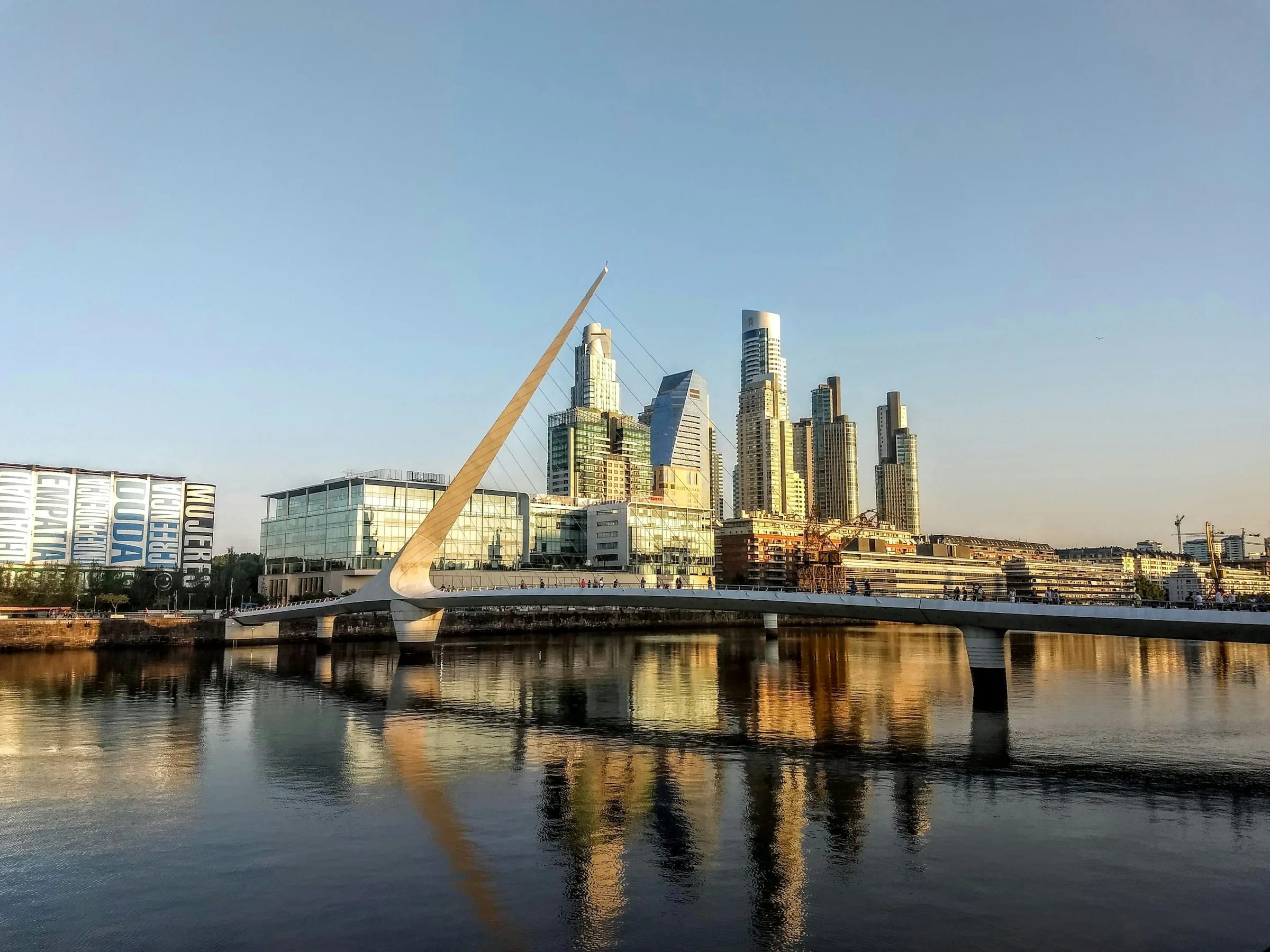 A white footbridge and the glass towers of Puerto Madero reflected in the water at golden hour in Buenos Aires.
