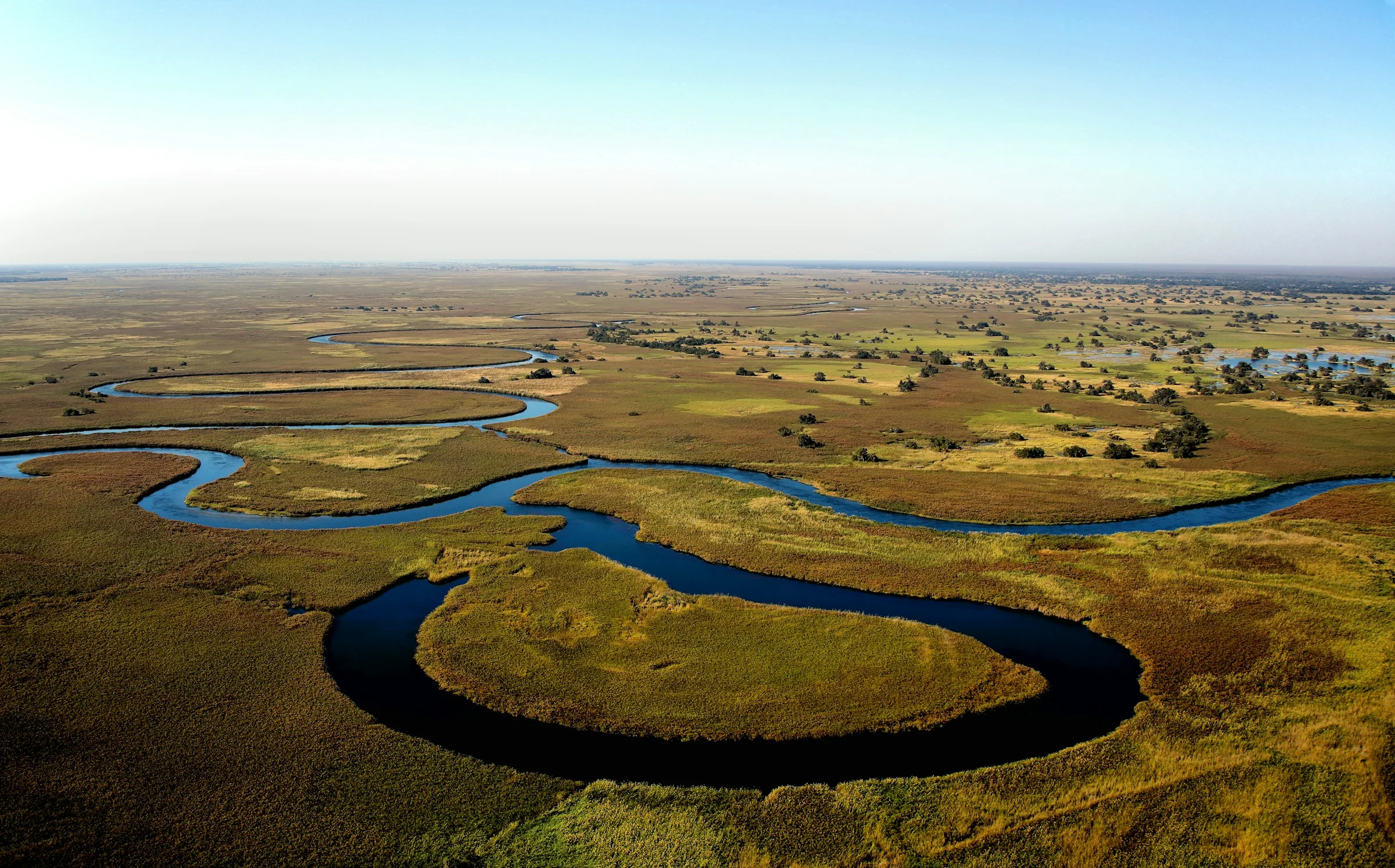 A river bending in a wide loop through green and golden floodplain, seen from the air under a clear sky.