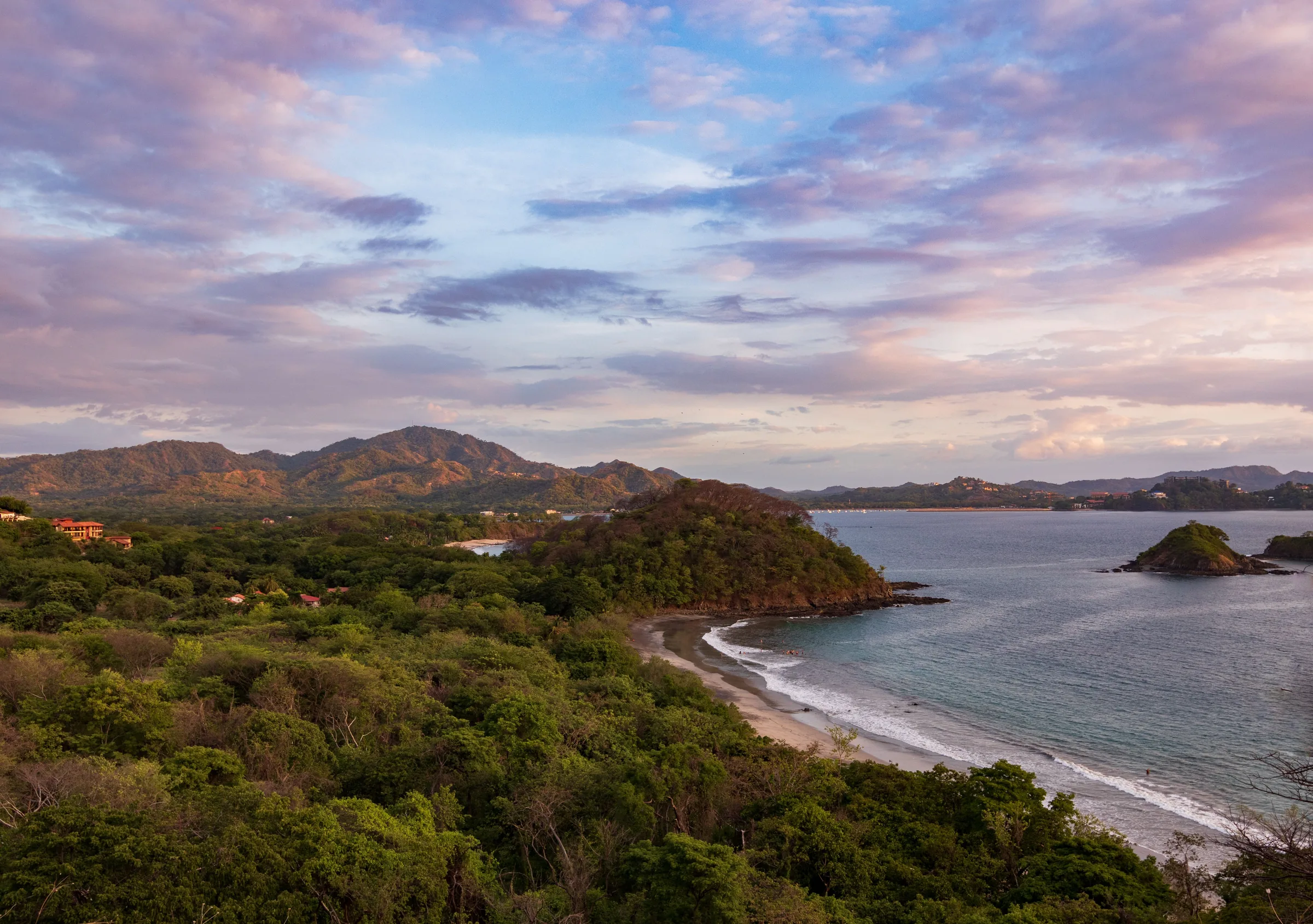 A forested headland, a sandy beach and a small rocky islet on the Guanacaste coast at dusk, with hills behind and pink clouds above.