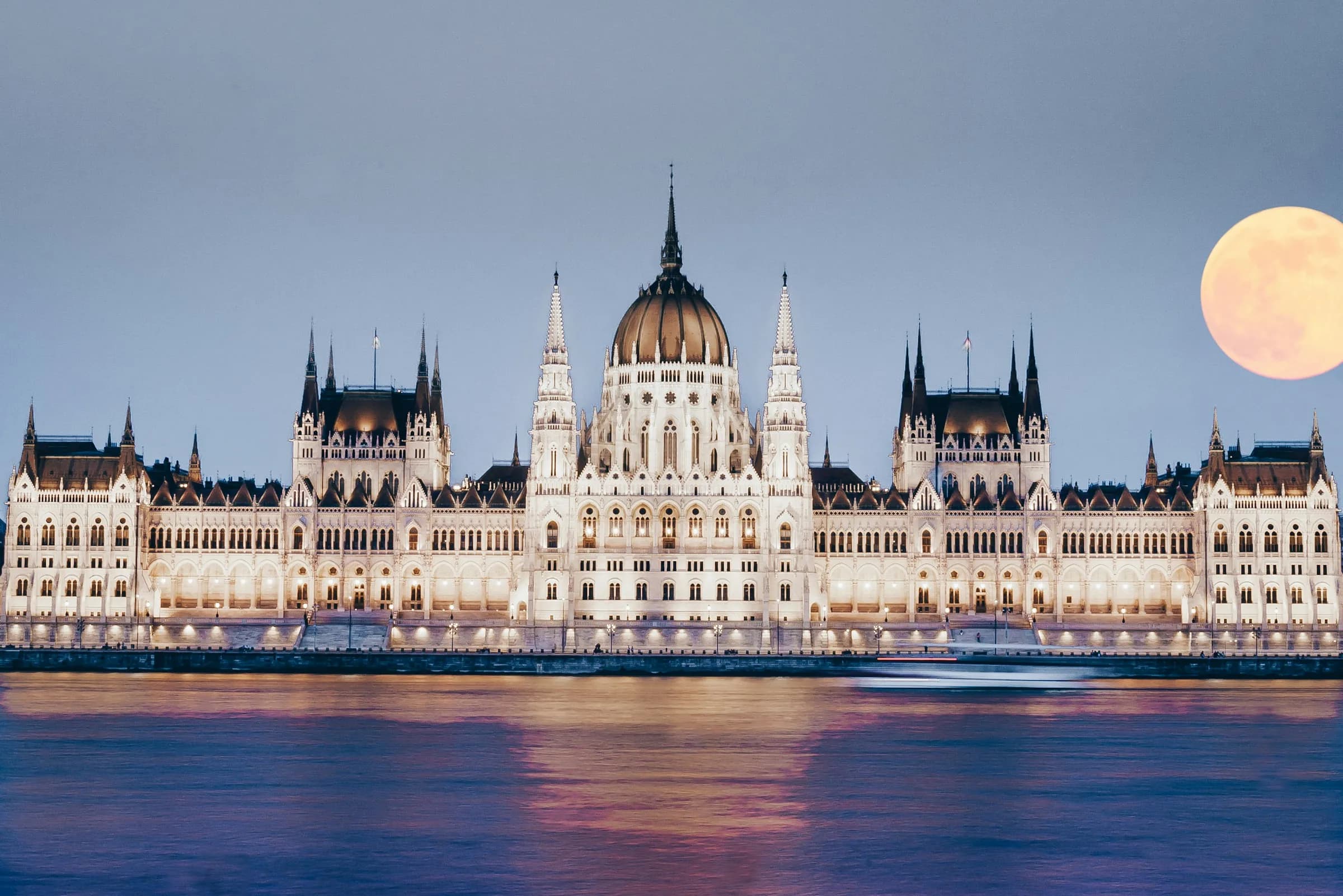 The Hungarian Parliament Building lit at dusk on the bank of the Danube in Budapest, with a full moon rising beside it.