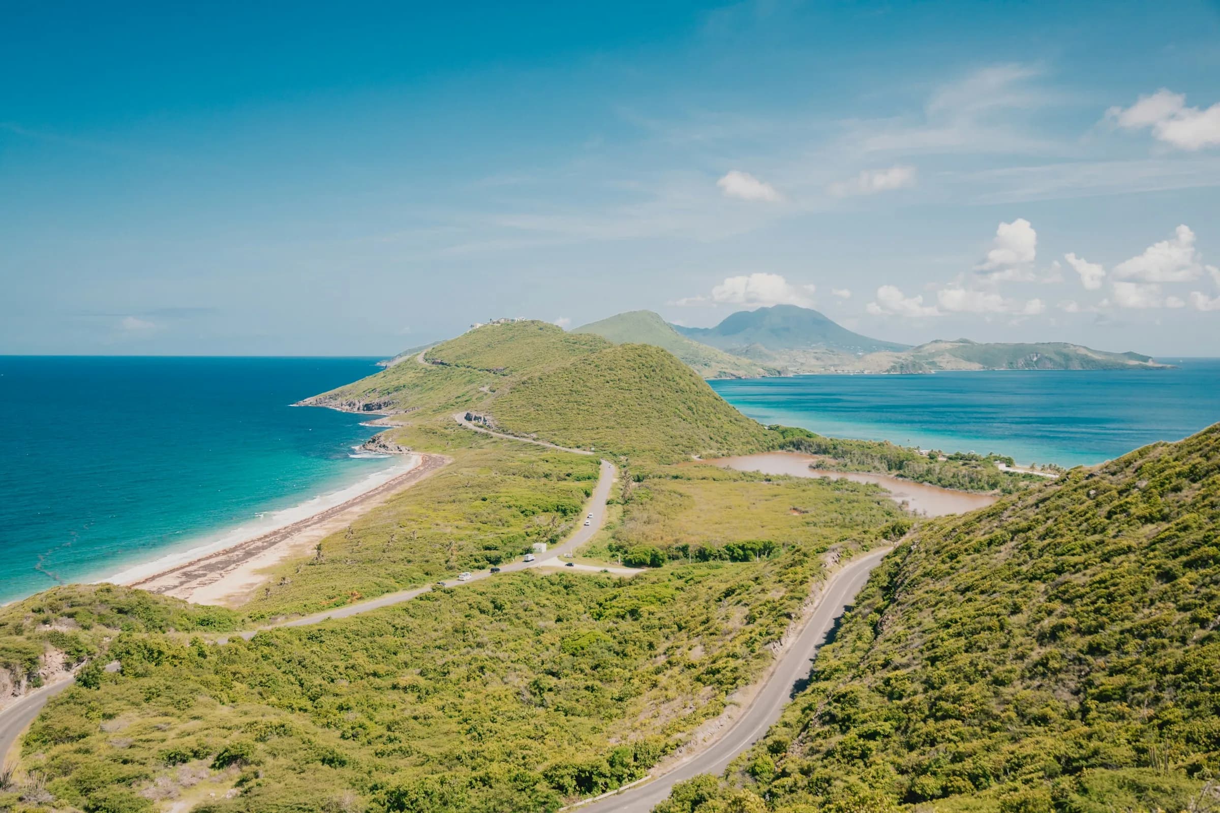 A road winding along a green headland between a turquoise sea and a sheltered bay, with a distant volcanic island under blue sky.