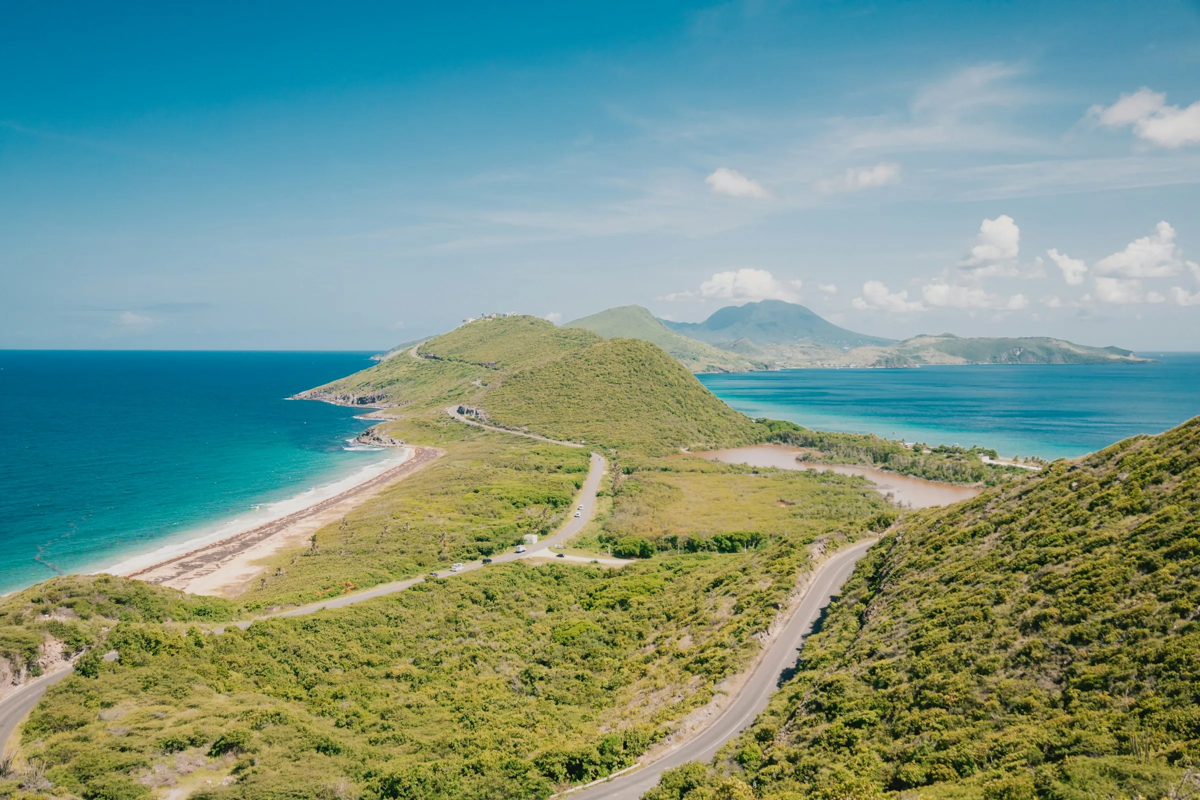 A road winding along a green headland between a turquoise sea and a sheltered bay, with a distant volcanic island under blue sky.