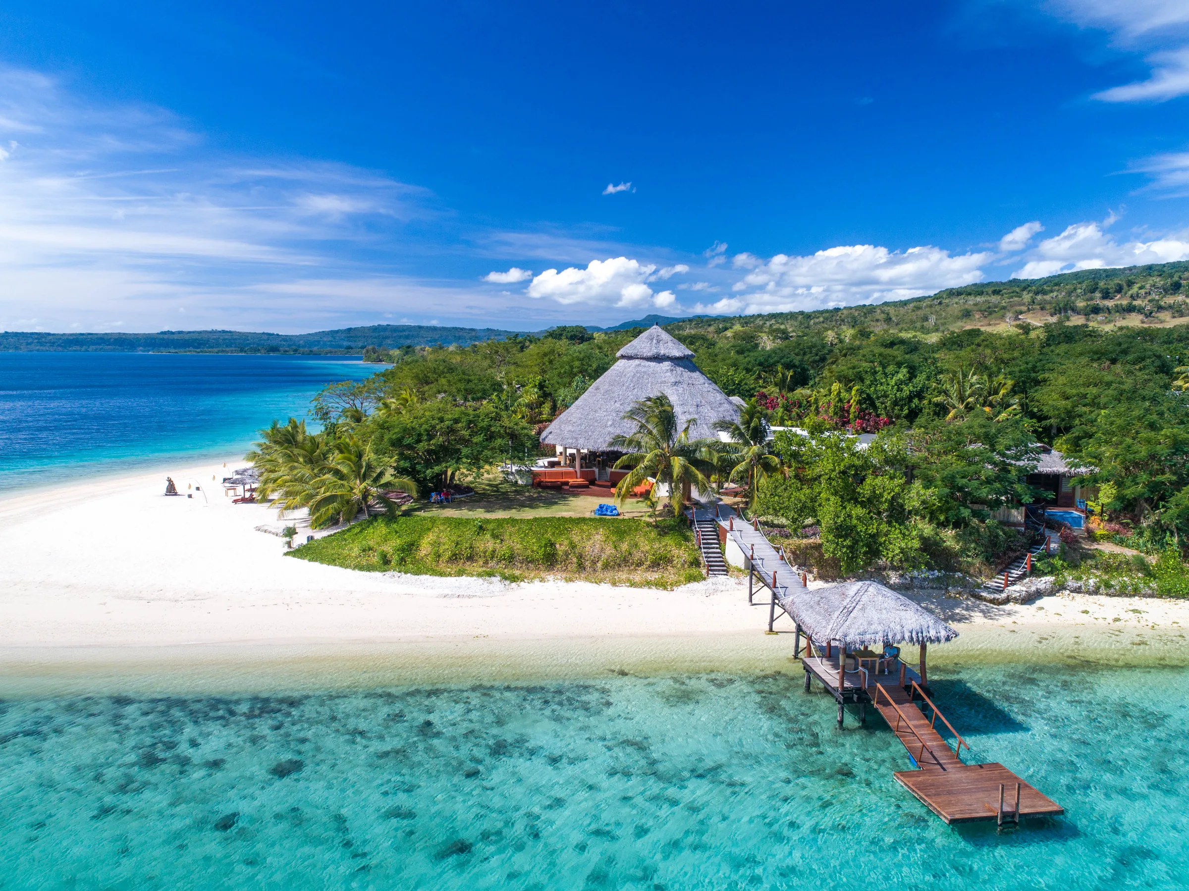 A thatched hut and a wooden jetty on a white sand beach beside a turquoise reef, with forested hills behind and a blue sky.