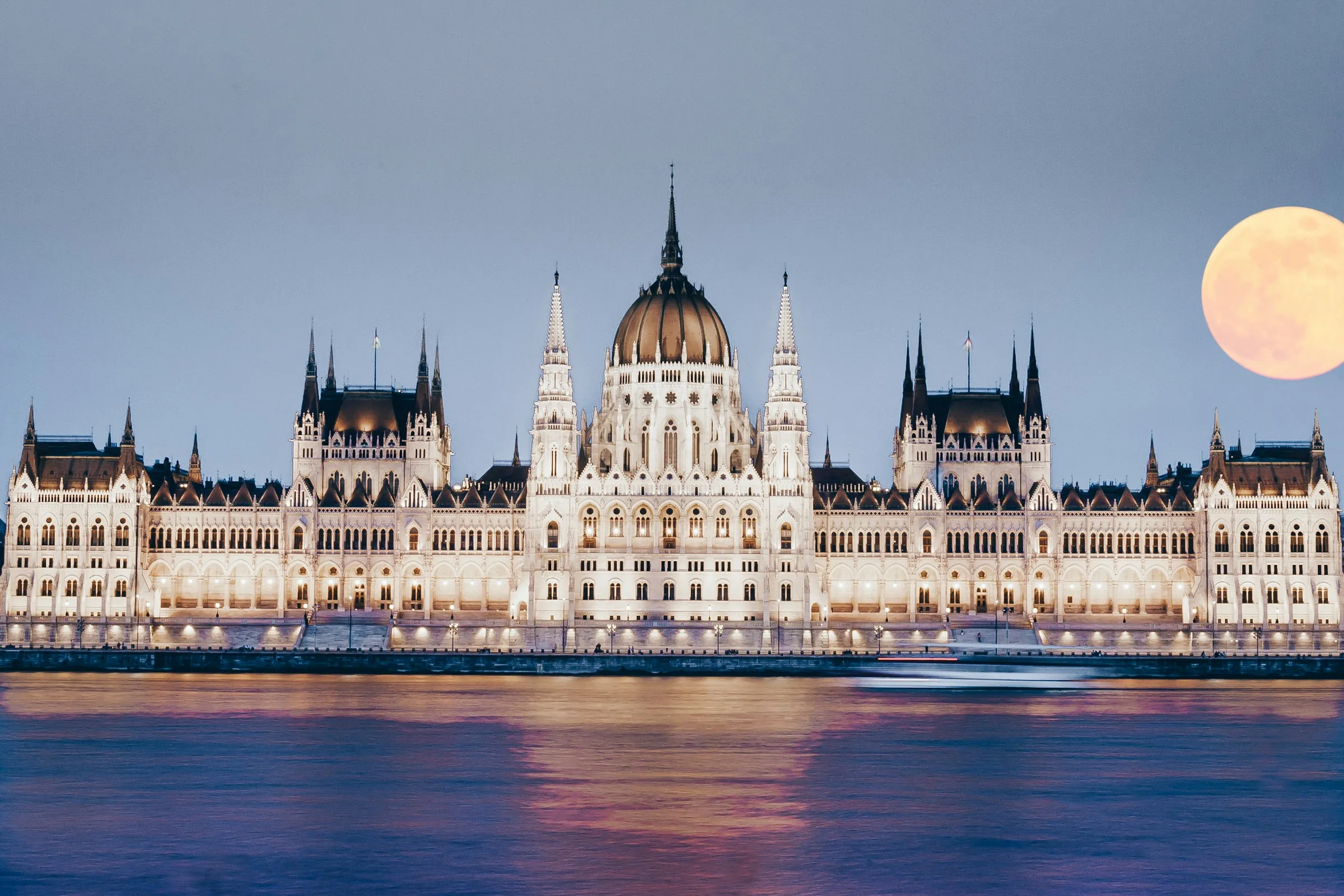 Das ungarische Parlamentsgebäude in der Abenddämmerung beleuchtet am Ufer der Donau in Budapest, daneben ein aufgehender Vollmond.